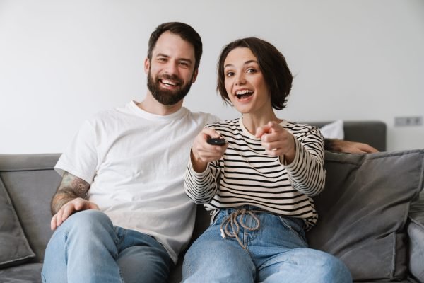 Happy young couple sitting on a couch at home Happy young couple sitting on a couch at home watching tv holding remote control